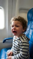 Toddler boy with curly brown hair crying on airplane seat wearing striped shirt during flight travel discomfort.