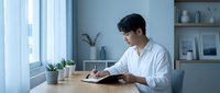 Young Asian male professional in white shirt working on digital tablet at wooden desk in modern office space with blue walls, potted plants, and natural light from window.