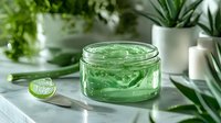 Fresh aloe vera gel in glass jar with cut leaf piece on white marble counter, surrounded by green plants and candles. Natural skincare concept.