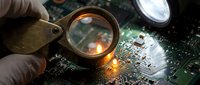 Technician examining computer circuit board with magnifying glass under bright light for electronic repair and diagnostics in professional workshop setting.