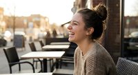 Young woman laughing joyfully at outdoor cafe terrace during golden hour sunset. Casual lifestyle moment of happiness and relaxation in urban setting.