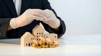 Businessman hands protecting wooden house models and gold coins symbolizing property investment security and real estate financial planning concept.