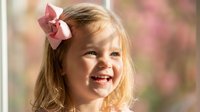 Happy blonde toddler girl with pink bow smiling joyfully outdoors in spring garden with soft floral background for children photography and family portraits.