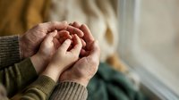 Senior woman hands clasped together in prayer or meditation pose wearing knitted sweater by window with soft natural light for wellness and spirituality concepts.
