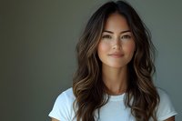 Young Asian woman with long wavy brown hair and natural makeup wearing white t-shirt smiling confidently against dark background. Professional headshot portrait.