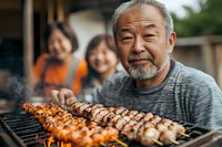 Senior Asian man grilling yakitori skewers at outdoor family barbecue, smiling while cooking traditional Japanese chicken kebabs over charcoal grill.