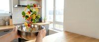 Fresh colorful vegetables falling into stainless steel bowl in modern kitchen. Healthy cooking and meal preparation concept for nutritious lifestyle.