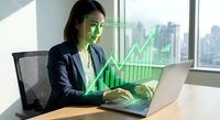Asian businesswoman working on laptop with holographic financial growth chart and green arrow overlay in modern office with city skyline view.