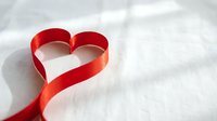 Red satin ribbon shaped into heart symbol on white fabric background for romantic occasions, weddings, valentines day celebrations and love concepts.