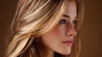 Young Caucasian woman with blonde highlighted hair and natural makeup in profile view against brown background, soft lighting creates warm atmosphere for beauty and fashion content.