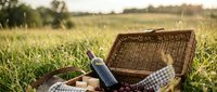 Wicker picnic basket with wine bottle, bread, and checkered cloth on sunny green grass field for outdoor dining and leisure activities.