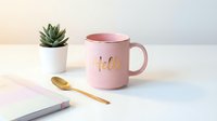Pink ceramic mug with gold Hello lettering beside a succulent plant, golden spoon, and pastel notebook on a white surface.