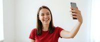Young Caucasian woman in red t-shirt taking selfie with smartphone, smiling happily at camera in bright modern interior for social media content.