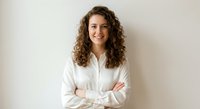 Professional young woman with curly brown hair in white shirt smiling confidently with crossed arms against neutral background for business content.