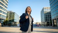 Young woman with long brown hair and light skin tone, wearing professional navy blazer and glasses, standing confidently in modern urban plaza surrounded by contemporary office buildings during daytime. Suitable for corporate, business, and professional lifestyle content.