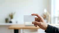 Person of medium skin tone holding blank white business card in modern bright office space with wooden desk and potted plants, suitable for corporate branding and professional networking concepts.