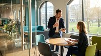Professional businessman and businesswoman having meeting discussion in modern office with large windows and city view during daytime collaboration.
