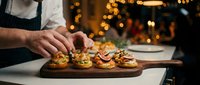 Chef preparing gourmet appetizers on wooden board in restaurant kitchen with warm bokeh lights creating festive culinary atmosphere for fine dining service.
