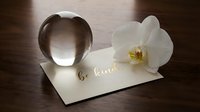 Crystal ball and white orchid on a cream card with gold foil be kind lettering, arranged on a dark wooden surface.