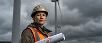 Confident male construction worker in hard hat and safety vest holding blueprints at industrial site with dramatic cloudy sky background.