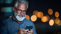 Mature bearded man in glasses using smartphone at night with glowing bokeh lights in background, expressing contentment and engagement.