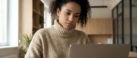 Young woman with dark curly hair and medium skin tone working on laptop in modern home office. Wearing beige turtleneck sweater. Suitable for business, technology, remote work, and lifestyle content.