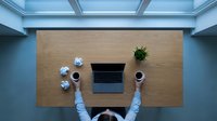 Overhead view of person at wooden desk with laptop, coffee cups, and plants in modern office workspace.
