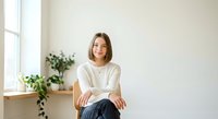 Young woman with long brown hair and fair complexion, wearing a white t-shirt and dark jeans, sitting by a bright window with potted plants. Suitable for lifestyle, wellness, and portrait use.