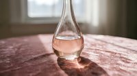 Elegant glass bottle with rose-colored liquid on wooden surface near window, casting soft shadows. Perfect for beverage, perfume, or cosmetic product marketing and packaging design.