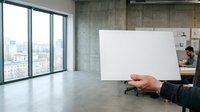 Businessman holding blank white card in modern office with floor-to-ceiling windows, concrete walls, and colleague working at desk in background. Corporate mockup for branding.