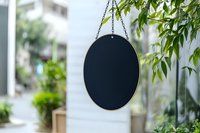 Oval black chalkboard sign hanging from chain against blurred garden background with green leaves, mockup for restaurant or cafe menu display outdoors.