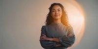 Young mixed-race woman with curly hair standing confidently with arms crossed against neutral background with golden sun halo effect.