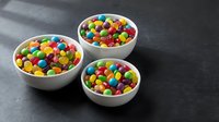 Colorful candy-coated chocolates in white ceramic bowls on dark slate surface. Sweet treats for parties, celebrations, and dessert displays.
