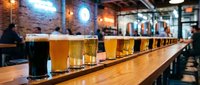 Craft beer flight with various golden and dark ales lined up on wooden bar counter in modern brewery taproom setting.