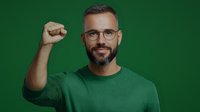 Confident Caucasian man with glasses and beard wearing green sweater showing raised fist gesture against green background, expressing determination and success.
