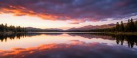 Serene lake at sunrise with vibrant orange and purple sky reflecting on calm water, surrounded by evergreen forest and distant mountains, peaceful nature landscape.