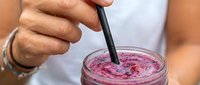 Person with medium skin tone stirring a pink smoothie or berry drink with a black straw, wearing a white shirt and bracelet. Closeup of refreshing beverage preparation.