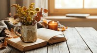 Cozy autumn still life with dried flowers in ceramic mug, open book and candle on wooden table by window creating warm peaceful reading atmosphere.