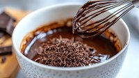 Melted dark chocolate dripping from whisk into white ceramic bowl with chocolate shavings, close-up shot showing glossy texture and rich brown color. Food preparation.