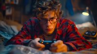 Young Caucasian man in glasses and red plaid shirt intensely focused on playing video games at night, lying on bed with game controller in atmospheric lighting.
