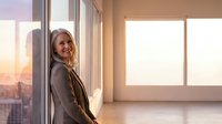 Professional blonde businesswoman smiling confidently in modern office with city skyline view during golden hour sunset, corporate success concept.