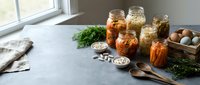 Homemade fermented vegetables in glass jars on kitchen counter with fresh herbs and wooden utensils for healthy meal preparation and food preservation.