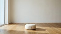 Minimalist interior with cream round ottoman on wooden floor in sunlit room with white walls and large window for modern home decor and furniture design.