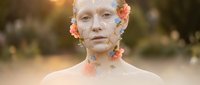 Young Caucasian woman with white clay face mask and colorful flowers adorning her face and neck in natural outdoor spa setting with warm golden sunlight.