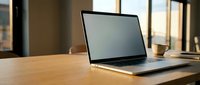 Laptop with blank screen on wooden desk in warm sunlit office, coffee cup in background, mockup ready for display presentation.