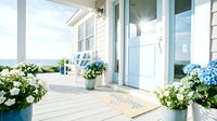 Sunny coastal cottage porch with blue door, welcome mat, white and blue hydrangeas, ocean view in background on a bright summer day.