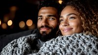 Young African American couple wrapped in cozy knit blanket sharing intimate moment outdoors at night with bokeh lights in background, expressing joy and romance.
