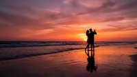 Romantic couple silhouette embracing on beach at sunset with dramatic orange and pink sky reflecting on wet sand and ocean waves.