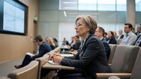 Professional blonde businesswoman in glasses attending corporate conference or seminar in modern meeting room with colleagues listening to presentation.