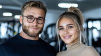 Young Caucasian couple smiling at camera in modern gym setting. Man wearing glasses and black sweater, woman with blonde hair in messy bun wearing beige turtleneck.